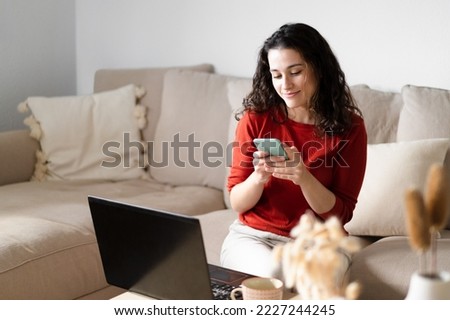 Young happy woman using the phone and  the computer sitting on the couch at home Royalty-Free Stock Photo #2227244245