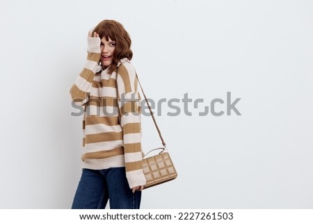 a lovely woman stands on a white background in a striped sweater and a bag on her shoulder, lowered her relaxed hand down, covering her eyes with the other