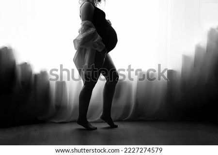 Black and white silhouette of a pregnant woman against the background of a window and curtains. Monochrome. Close up. Pregnancy concept.