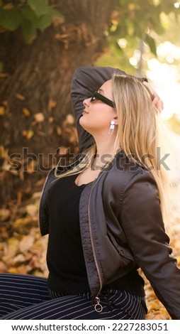 Blonde girl wearing glasses and squatting down posing for a picture in a beatiful fall scenery during a sunny day.