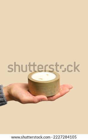 A woman's hand holds a decorative candle in a golden pot on a beige background, minimal composition