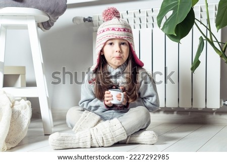 Grimacing little girl in hat, socks and pullover drink sour medicine syrup and sit near heating convector, radiator, next to houseplant. Seasonal sick. Weak throat, immune system in cold winter Royalty-Free Stock Photo #2227292985
