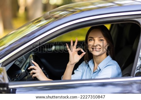 Attractive girl is sitting in a modern car. She is looking through window and showing okay sign. The lady is smiling with joy
