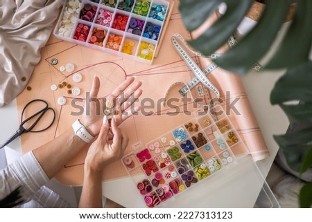 Woman tailor choosing sew button round heart shape creating clothes garment at studio top view closeup. Female fashion designer dressmaker trying material for clothing repair enjoy art hobby work Royalty-Free Stock Photo #2227313123