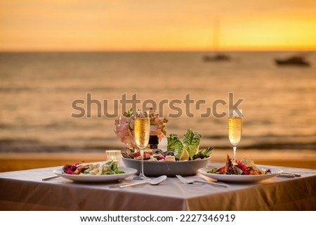 Romantic sunset dinner on beach. Table honeymoon set for two with luxurious food, glasses of champagne drinks in restaurant with sea view and yachts on background. Summer vacation or wedding concept. Royalty-Free Stock Photo #2227346919