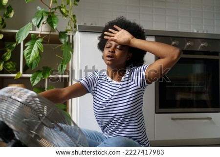 Sweaty overheated young African woman using electric fan ventilator, black girl suffering without air conditioner at home, touching forehead feeling unwell, staying indoors during extreme summer heat Royalty-Free Stock Photo #2227417983