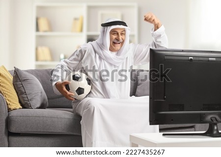 Mature arab man in a robe holding a football and cheering in front of tv at home Royalty-Free Stock Photo #2227435267