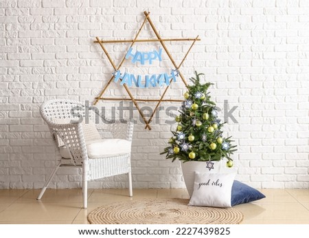 David star with text HAPPY HANUKKAH, armchair and fir tree near white brick wall Royalty-Free Stock Photo #2227439825