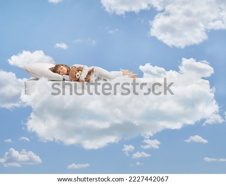 Little girl in pajamas holding a teddy bear and sleeping on clouds Royalty-Free Stock Photo #2227442067