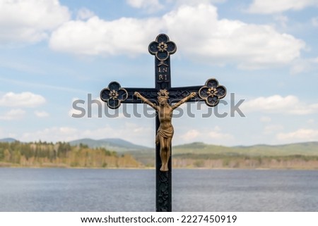 Crucifix - statue of Jesus Christ on a cross with the inscription INRI, by the pond, in the background landscape with water, hills and blue sky with white clouds