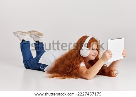 Watching cartoons. Cute redheaded little girl wearing tee and jeans using tablet isolated over white studio background. Remote online education, kids emotion, facial expressions, fashion