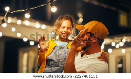 Cinematic Shot of a Father Holding His Small Daughter in His Arms, Helping Her to Fix a Lightbulb in a Backyard Lights Installation. Father and Daughter High Five and Celebrate Successful Repair. Royalty-Free Stock Photo #2227481151