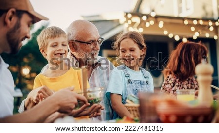 Happy Senior Grandfather Talking and Having Fun with His Grandchildren, Holding Them on Lap at a Outdoors Dinner with Food and Drinks. Adults at a Garden Party Together with Kids. Royalty-Free Stock Photo #2227481315