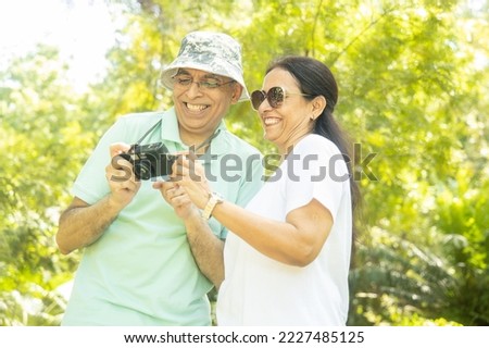Happy senior indian couple tourist holding camera taking photos and view them together at summer park. Mature people travel and holidays.