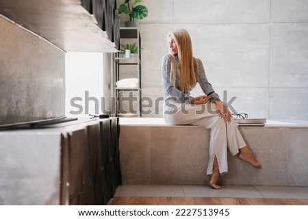 Frustrated  blonde fit young woman in white pants and shirt holding phone sitting on bath at bathroom looks aside with calm face. Beautiful luxury Swedish female alone at home. Luxury lifestyle. Royalty-Free Stock Photo #2227513945