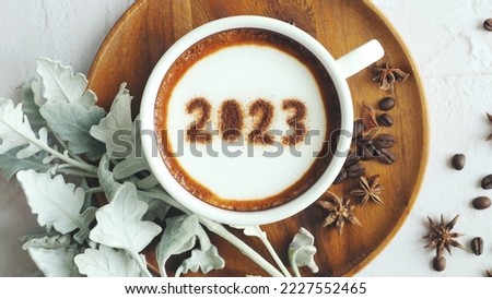 Happy New Year 2023 theme coffee cup with number 2023 over frothy surface served on wooden saucer and white cement background with coffee beans, star anise, silvery gray foliage of dusty miller plant.