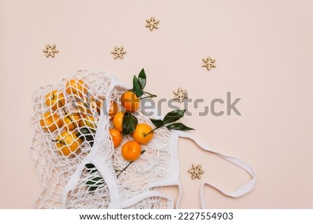 Top view of a white grocery eco-mesh cotton bag with fresh orange tangerines on a beige pastel background and scattered golden snowflakes. Buying tangerines for the new year.