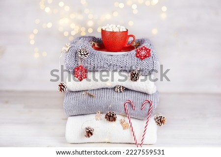 A stack of folded knitted sweaters, a red cup of cocoa with marshmallows, a garland with Christmas decorations on a background of bokeh lights. Cozy homely atmosphere of the Christmas holiday. 