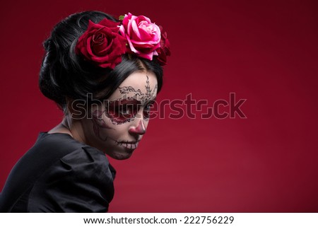 Close-up portrait of young girl in black dress with Calaveras makeup and a red flower in her black hair looking aside isolated on red background with copy place
