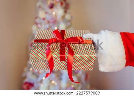 Santa Claus's hand in a white glove holds a gift box on the background of a decorated light Christmas tree