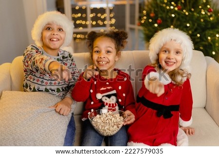 A children in Santa hats watch movies at home with with a big bowl of popcorn on Christmas day pointing at the screen