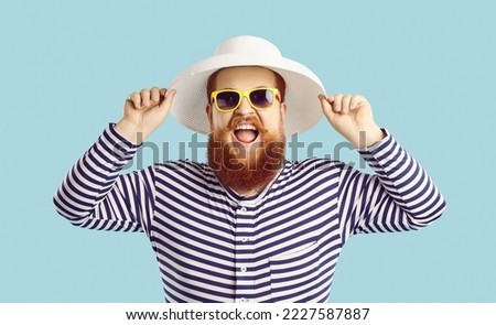 Summer time. Funny excited chubby man with white beach hat isolated on pastel light blue background. Close up portrait of crazy redhead bearded man in striped clothes and sunglasses laughing out loud.