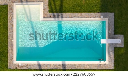 Aerial view of a rectangular swimming pool with diving board, belonging to a large villa. The water is transparent and through the blue you can see the steps. Around the water there is a stone floor. Royalty-Free Stock Photo #2227603559
