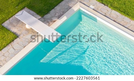 Aerial view of a rectangular swimming pool with diving board, belonging to a large villa. The water is transparent and through the blue you can see the steps. Around the water there is a stone floor. Royalty-Free Stock Photo #2227603561