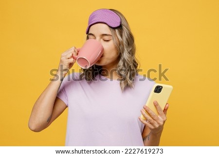 Calm young woman wears purple pyjamas jam sleep eye mask rest relax at home hold use mobile cell phone cup drink tea isolated on plain yellow background studio portrait. Good mood night nap concept