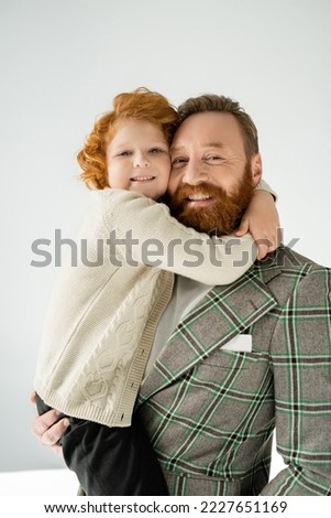 Red haired boy hugging stylish parent and looking at camera on grey background