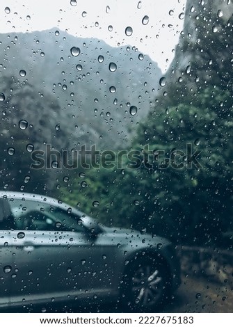 Raindrops on the clean car glass image with silver color car on the background. Deep autumn cold rainy foggy day photo on the mountain parking spot. Weather conditions and modern cars traffic concept.