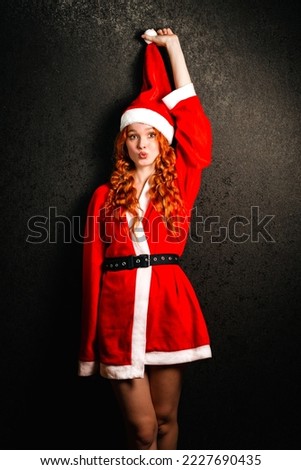 A happy young girl in a Santa Claus costume smiles on a black background. Portrait of a red-haired woman in a red cap laughing on a dark background. The concept of Christmas and New Year.