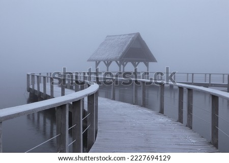 Scenic lake side view with wooden jetty in dense fog in winter. Royalty-Free Stock Photo #2227694129