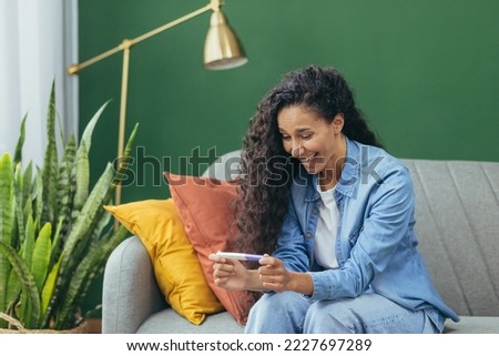 Happy young beautiful hispanic woman sitting on sofa at home, holding home pregnancy test. Satisfied with the positive result.