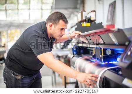 In the printing house, an experienced technician works on a UV printer. Production work. Check the print quality. Royalty-Free Stock Photo #2227705167