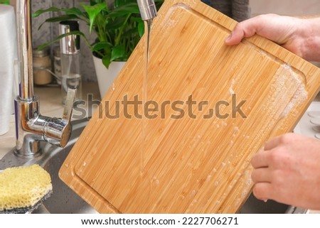 A man washes a wooden bamboo cutting board in the kitchen sink under running water. Gentle hand washing of a wooden cutting board. Cleaning dirty kitchen wood products. Royalty-Free Stock Photo #2227706271