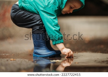 A small child boy wearing blue rubber boots and a jacket jumping through puddles and playing outside on a rainy day. Having fun outdoors during a bad weather. Hands in a water. Copy space. Royalty-Free Stock Photo #2227722415