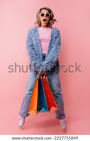 Full length playful young blonde caucasian woman with open mouth holds packages on pink background. Girl wears sunglasses, fur coat and jeans. Shopping concept