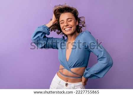 Joyful young caucasian woman closing eyes smiling broadly having fun on purple background. Blonde wears blue blouse and pants. Mood, lifestyle, concept.