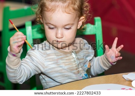 Toddler girl during an art project. Kid holds paintbrush. Baby paints with brush on a paper. A girl paints a picture