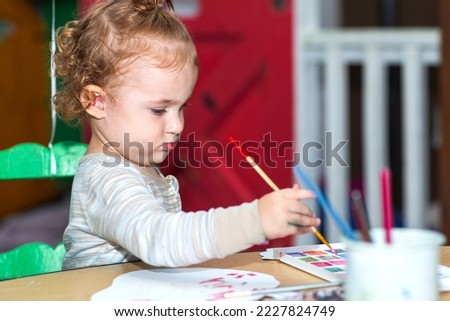 Toddler girl during an art project. Kid holds paintbrush. Baby paints with brush on a paper. A girl paints a picture