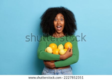 Amazed dark skinned curly woman holds a bunch of fresh oranges and lemons, looks excited, wears green cardigan and blue jeans, isolated over blue background 