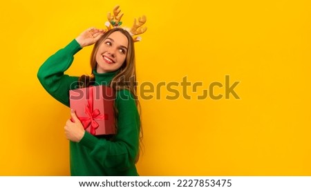 Happy smiling woman in Christmas reindeer antlers headwear and green sweater holding pink present gift box and looking up on a yellow background with copy space.

New Year and Christmas concept.