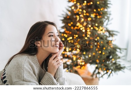 Portrait of young smiling asian woman with dark long hair in cozy sweater in room with Christmas tree