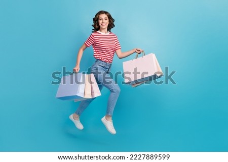 Full length photo of sweet cute lady wear striped t-shirt jumping high holding bargains running isolated blue color background Royalty-Free Stock Photo #2227889599