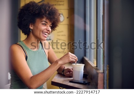 Female Customer In Window Of Cafe Stirring Hot Drink In Takeaway Cup With Wooden Stirrer Royalty-Free Stock Photo #2227914717