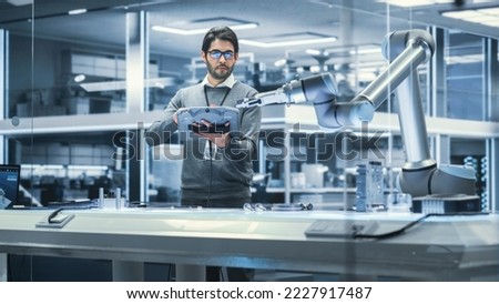 Robotics Engineer Researching and Developing a Technological Robot Arm Project, Standing with Tablet Computer in Scientific Technology Lab. Young Male Working on an AI Robotic Arm. Royalty-Free Stock Photo #2227917487