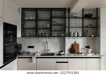 Interior design of stylish kitchen with gllas, gray gabinets, black oven, coffee machine, cutting board, bowl with avocado, silver tap, kitchen towel and personal accessories. Home decor. Template. 
