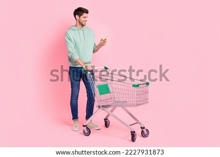 Full length photo of positive stylish man hold pushcart read list buy products supermarket empty space isolated on pink color background Royalty-Free Stock Photo #2227931873