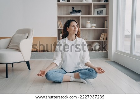 Peaceful hispanic woman practicing yoga sitting in padmasana, lotus pose on floor in living room. Calm female meditating, doing breathing exercises at home. Stress relief, healthy lifestyle concept. Royalty-Free Stock Photo #2227937235